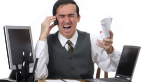 male surrounded by screens with hand to head and eyes closed show how work can cause stress